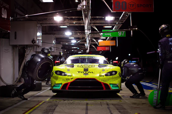 Aston Martin Vantage GTE race car in pit box during night pit stop at Le Mans, mechanics changing tyres under bright garage lights, endurance racing strategy and precision in FIA WEC