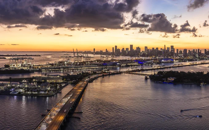 Aerial shot of Miami at dusk. Water reflects golden sunlight. A long bridge crowded with cars leads to a skyline filled with lit buildings. Cranes populate the dock as boats cruise through the water.