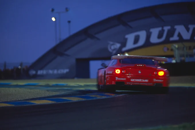 Night shot of red Ferrari 550 GTS Maranello at Circuit La Sarthe during  24 Hours of Le Mans 2002, Prodrive built race car takes a corner with glowing brake lights, and  Dunlop bridge in the background.
