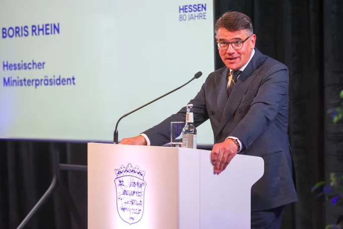 Ein Mann mit Brille steht an einem Rednerpult und hält eine Rede bei einer Veranstaltung, wie auf dem Bildschirm hinter ihm zu sehen ist.