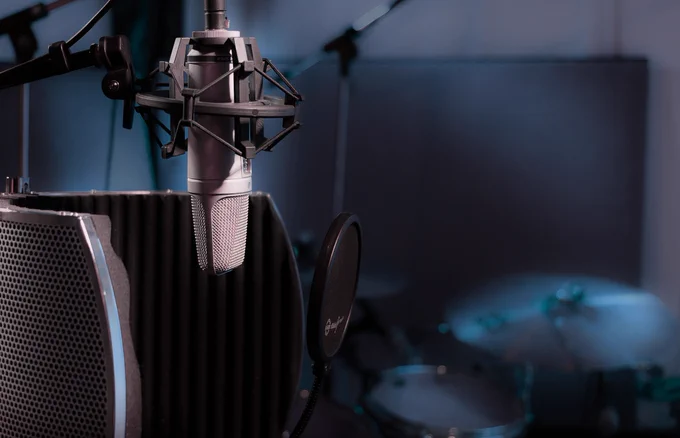 Professional recording studio setup featuring a condenser microphone with shock mount and pop filter in front of a sound isolation shield, with a blurred drum kit in the background under soft blue lighting.