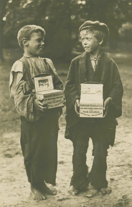 A photograph of two small boys from the late Victorian/early twentieth century looking towards each other. They are dressed in clothing typical of those with little income in the period. They are both smiling and holding collection boxes. A wooded scene behind. 