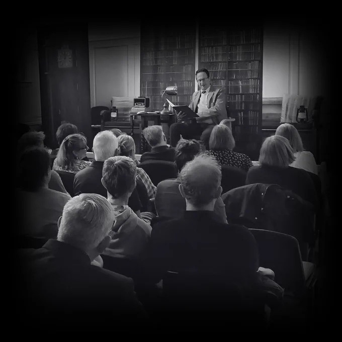 A room of people listening to The Librarian tell a story in an old building. He has bookcases and a Grandfather clock behind him and a table with a lamp next to him.