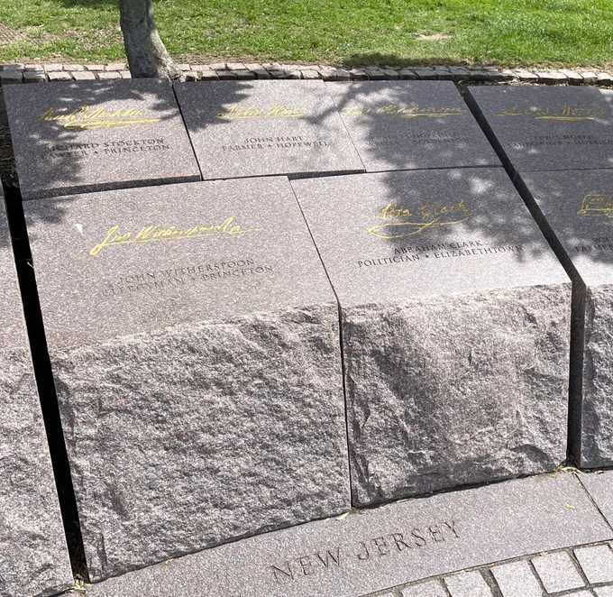 Five reddish gray stone blocks with names etched into the tops and gold signatures on them and "New Jersey" written at the base stand as a section of a low memorial wall.