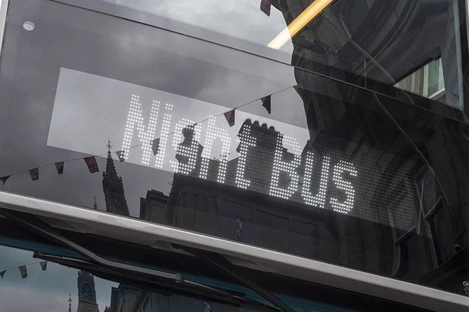 Night BUS displayed in white LED dots on a bus's digital sign, with reflections of buildings and flags.