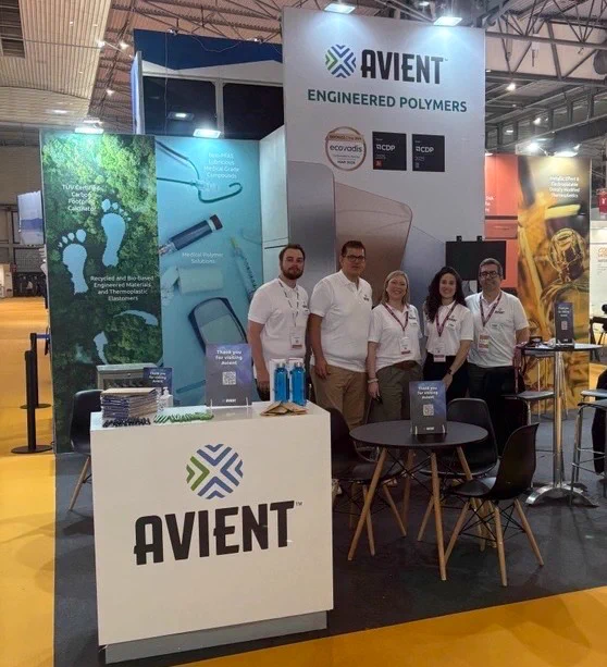 A group of people stands in front of an Avient booth at an event. The booth features displays about engineered polymers, with eco-themed graphics and information. A table with chairs is nearby, and the booth has visible signage with the Avient logo.