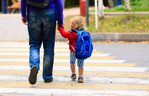 Adult and young child holding hands while crossing a pedestrian crosswalk on a sunny day.