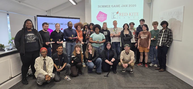 A group of people standing in a room, some holding small trophies. In the background are presentation slides with "Summer Game Jam 2026," "Red Kite Games," and "Leeds Trinity University" logos.