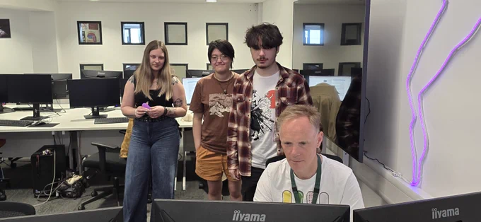 A group of four people in a computer lab, with three standing behind one who is seated at a desk working on a computer. The room has several monitors and a white board.