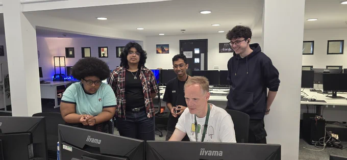 A group of five people gathered around and looking at computer monitors. The setting appears to be an office or computer lab with several desks and screens. They seem focused and engaged in the activity on the screens.