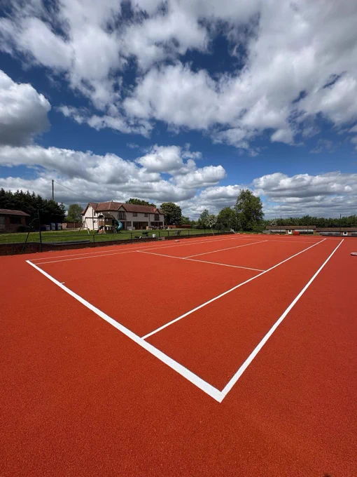 Empty red clay tennis court with white lines under a partly cloudy blue sky near residential houses.