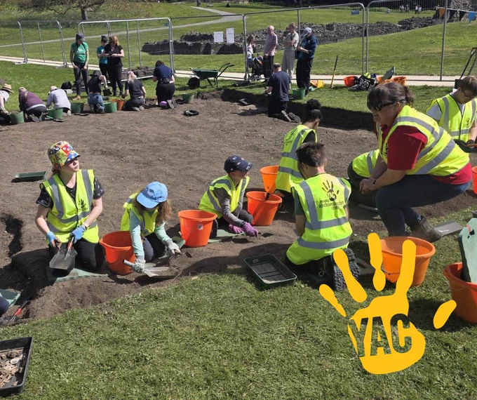 A group of children and adults working on an archaeological dig on a sunny day.