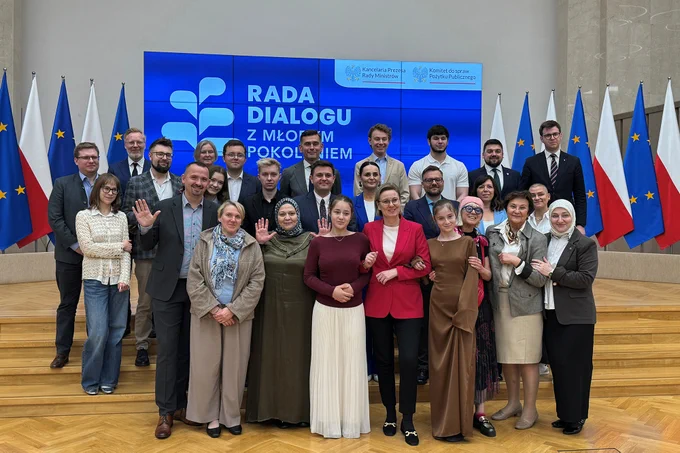 Zdjęcie grupowe. Uczestnicy wydarzenia pozują do wspólnego zdjęcia. W pierwszym rzędzie widoczna między innymi Sekretarz Stanu Adriana Porowska. W tle widoczny ekran, na którym wyświetlone jest logo Rady Dialogu z Młodym Pokoleniem. Po obu stronach ekranu stoją flagi Polski i Unii Europejskiej.