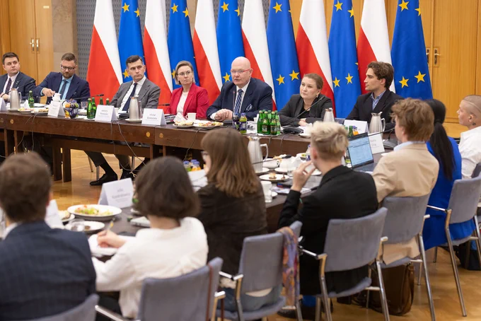 Szeroki kadr, widok obejmuje część stołu konferencyjnego, który ustawiony jest w kształcie litery „U”. Przy stole prezydialnym widoczni między innymi dwaj Współprzewodniczący Rady – Pan Hubert Taładaj i Pan Piotr Stec, a także Sekretarz Stanu Adriana Porowska. W tle widoczne flagi Polki i Unii Europejskiej. Na pierwszym planie widoczni uczestnicy spotkania, siedzący przy stole, tyłem do obiektywu.