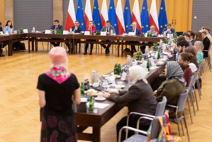 Szeroki kadr, widok obejmuje salę i stół konferencyjny ustawiony w kształcie litery „U”. Przy stole prezydialnym widoczni między innymi dwaj Współprzewodniczący Rady – Pan Hubert Taładaj i Pan Piotr Stec, a także Sekretarz Stanu Adriana Porowska. W tle widoczne flagi Polki i Unii Europejskiej. Na pierwszym planie rozmyte sylwetki uczestników.