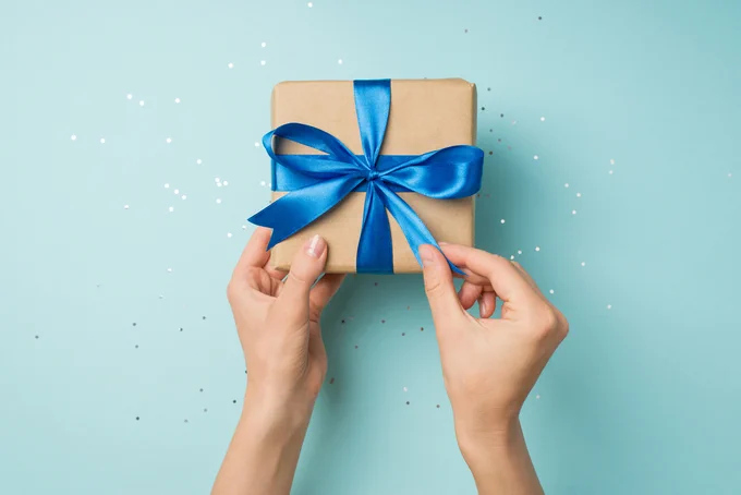 Hands tying a shiny blue ribbon around a small brown gift box on a light blue background with scattered white confetti.