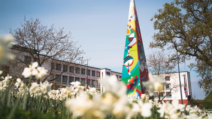 Colorful tall sculpture with abstract designs stands in front of a modern building surrounded by blooming white flowers and trees.