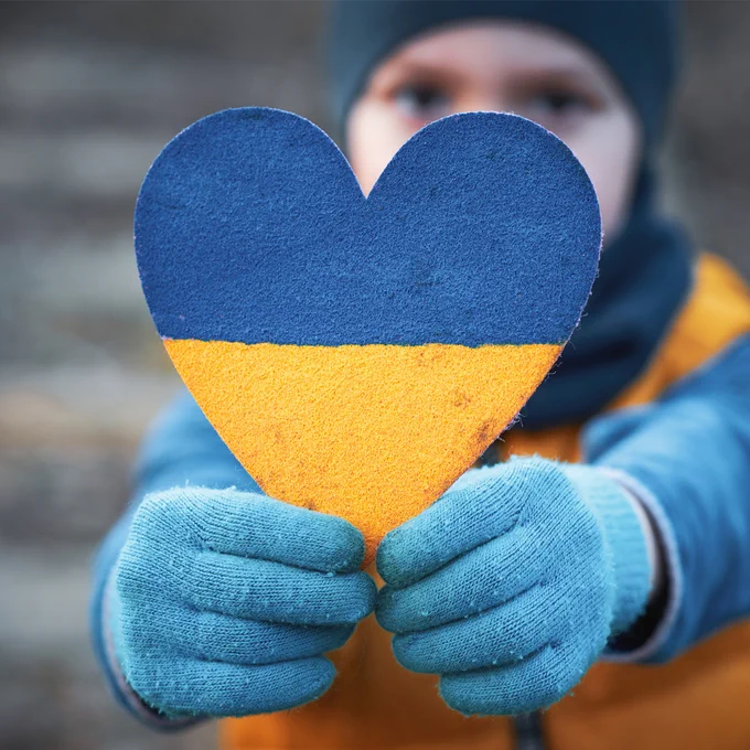 Person wearing blue gloves and winter clothing holds a heart-shaped cutout painted in the blue and yellow colours of the Ukrainian flag toward the camera. The heart is in sharp focus, while the person's face and background are softly blurred.