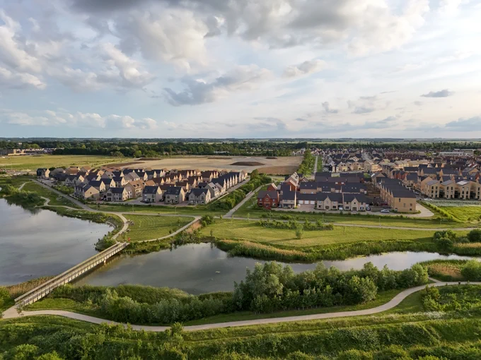 Aerial view of a modern housing development in Northstowe, with rows of new homes surrounded by green open space, a lake with a footbridge in the foreground, and countryside stretching into the distance under a partly cloudy sky.