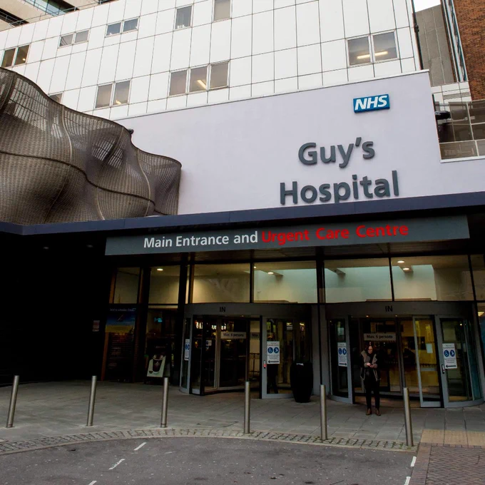 Entrance to Guy's Hospital with NHS signage and urgent care center highlighted in red above glass doors.