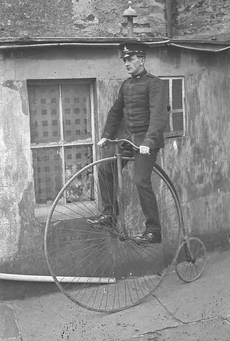 A black and white archive photograph of a man in uniform riding a penny-farthing with a large front wheel and small back wheel