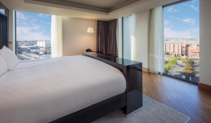 Modern hotel bedroom with a large white bed and dark wood frame beside floor-to-ceiling windows. Light curtains frame views of a waterfront cityscape with buildings, roads, and water under a bright sky.