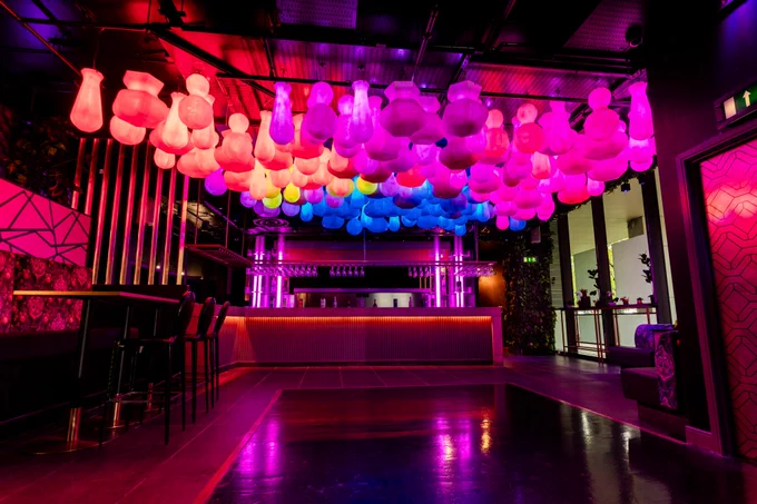 Stylish bar interior lit with vivid pink, purple, and blue lighting, featuring a ceiling installation of glowing, bulb-shaped lights. A bar counter with stools sits beneath the lights, surrounded by sleek, contemporary décor.