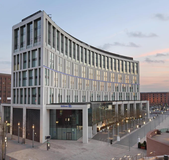 Curved modern hotel building with a white and glass façade, featuring multiple rows of windows and the Hilton logo near the entrance. The structure sits beside a wide, open plaza with paving, streetlights, and a few trees, under a soft evening sky with light clouds.