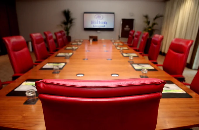 Conference room with a long wooden table surrounded by red leather chairs, each place set with notepads and glasses. A screen at the far end displays “Hilton Liverpool,” with plants and soft lighting in the background.