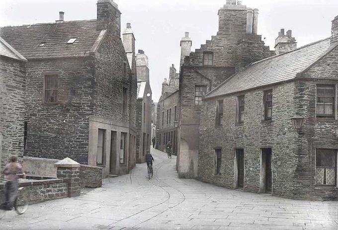 A colourised Tom Kent photograph of Dundas Street in Stromness taken around 1900. Several people are cycling on the street.