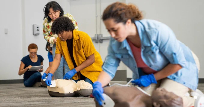 A group of individuals practice CPR and the use of automated external defibrillators (AEDs) on manikins in a classroom setting. One person in the background takes notes on a tablet. They are all focused on the training exercise.