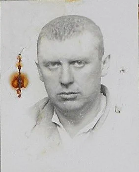 Black and white passport-style photo of a man with short hair, staring directly at the camera, wearing a light-colored shirt. The image has visible damage, including stains and holes.
