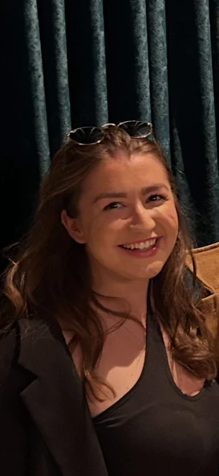 a picture taken of rachel who is sat down against dark curtains. She has sunglasses propped on her head, is wearing black, and  smiles at the camera