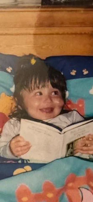 Rachel as a child, in bed reading a story. She has a raised strawberry birthmark on her cheek and has a 90s fringe and high ponytail