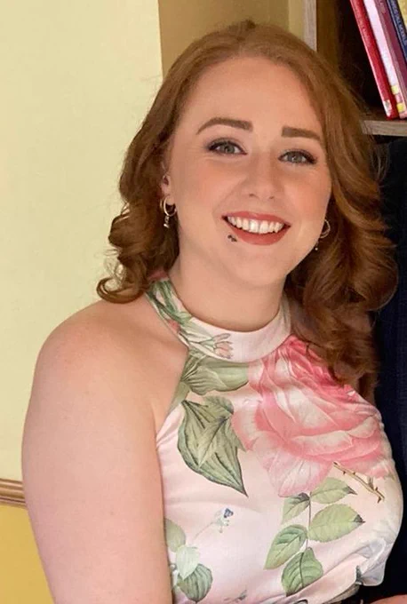 Niamh Murphy, with shoulder-length curly hair wearing a sleeveless floral top with pink and green leaves, standing indoors near a bookshelf.
