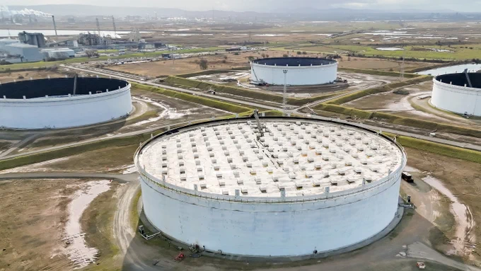A wide view of several large, circular oil storage tanks situated in an open, industrial landscape. The tanks are surrounded by grassy areas and connected by roads. 