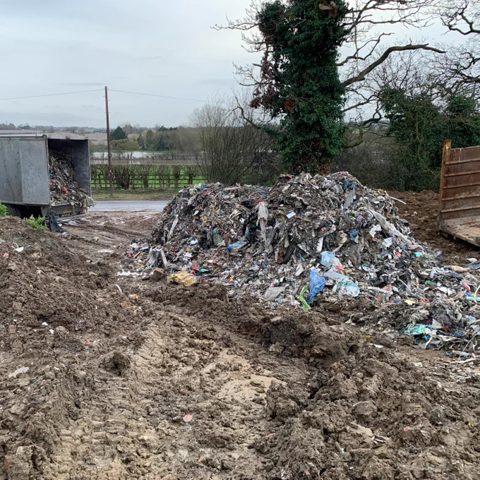 A large pile of mixed waste has been unloaded onto a muddy field. The adjacent road is open to the field where a lorry trailer full of more mixed waste can be seen with the doors open. 