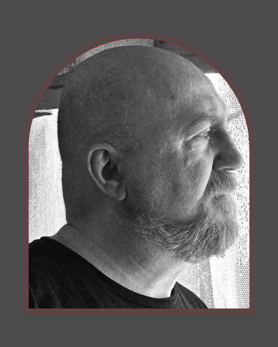 Black-and-white photo of a white man in profile, looking toward the right of the image. He is bald and has a beard and is standing in front of a lacy curtain
