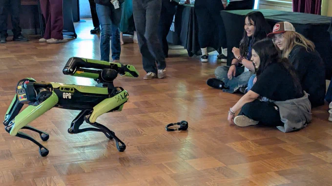 A small green and black 4-legged robot moves across a wooden floor while 3 people sit nearby watching. A pair of headphones rests on the floor in front of the robot, and tables and other attendees are visible in the background. 