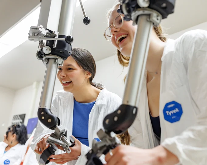 Two people in white lab coats closely examine and adjust mechanical laboratory equipment made up of metal arms and knobs. 
