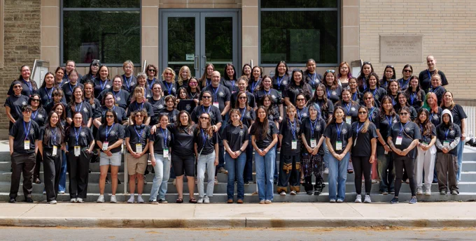 About 65 people, wearing matching dark t-shirts bearing the STEM Workshop for Indigenous Girls  logo, stand on steps outside a building at McMaster University. 