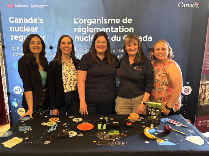 Five people at a Canadian Nuclear Safety Commission booth at an indoor event pose for a photo. Behind them is a bilingual backdrop that bears the words “Canada’s nuclear regulator / L’organisme de réglementation nucléaire du Canada” and the Government of Canada logo. The table in front displays various items, including radiation detection instruments, information materials, cards, pens, and everyday objects such as a banana and an apple for demonstration purposes.