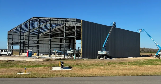 A large steel structure under construction with partial walls, featuring two blue cranes. The scene is set against a clear blue sky and grassy surroundings.