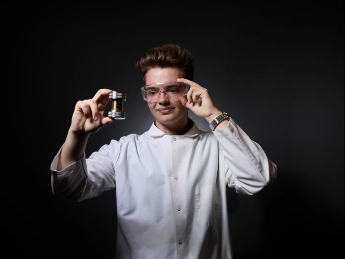 A person in a white lab coat and safety goggles holds up a small cylindrical object against a dark background, appearing focused on the item.