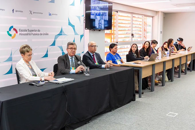 Imagem de mesa retangular preta com participantes homens e mulheres de uma reunião, fundo branco com a logo do ESMPU. Foto: Daniel Moutinho/Comunicação MPF.