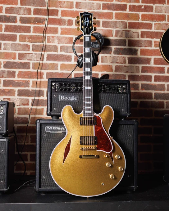 A gold sparkle Gibson Custom ES-355 electric guitar is displayed in front of a brick wall. Behind it, there are Boogie and Mesa guitar amplifiers. The guitar has a polished body with knobs and pickups visible. Headphones hang on a hook above the amplifiers.