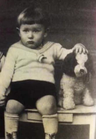 A black and white historical photo of a young boy sitting on a small stool next to a plush toy dog. The child has short hair and wears a light sweater and dark shorts, with socks pulled up to mid-calf, looking directly at the camera with a neutral expression. The toy dog beside the child appears fluffy and is sitting upright.
