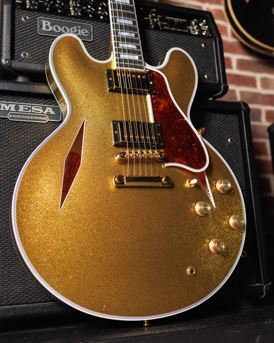 A glittery gold electric Gibson ES guitar leaning against a Mesa Boogie amplifier. The guitar has a semi-hollow body, two humbucker pickups, and multiple control knobs. The amplifier sits atop a speaker cabinet, with a brick wall backdrop.