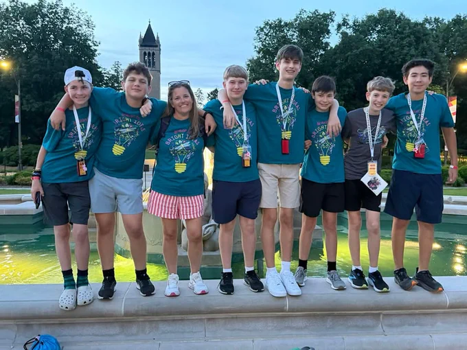 Group of seven young people standing arm in arm on a fountain ledge at dusk, wearing matching event shirts and lanyards.