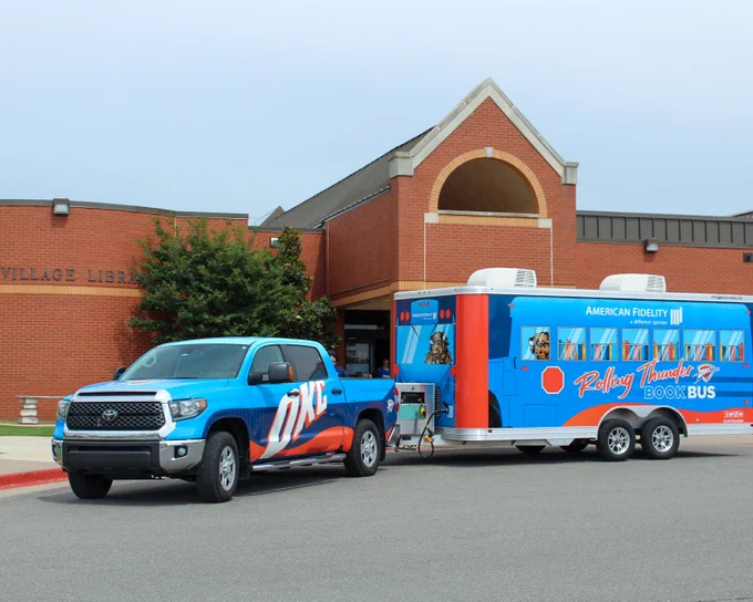Blue OKC pickup truck towing a blue and orange trailer for the Rolling Thunder Book Bus parked outside The Village Library.